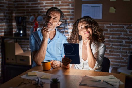 Photo for Middle age hispanic couple using touchpad sitting on the table at night with hand on chin thinking about question, pensive expression. smiling with thoughtful face. doubt concept. - Royalty Free Image