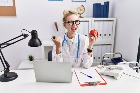 Foto de Young doctor woman working at dietitian clinic pointing thumb up to the side smiling happy with open mouth - Imagen libre de derechos