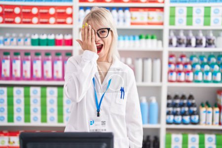 Téléchargez les photos : Jeune femme caucasienne travaillant à la pharmacie pharmacie couvrant un oeil avec la main, sourire confiant sur le visage et l'émotion surprise. - en image libre de droit