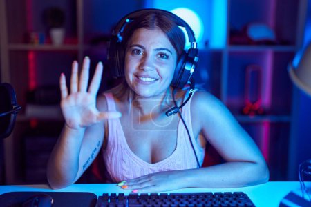 Foto de Young blonde woman playing video games wearing headphones showing and pointing up with fingers number five while smiling confident and happy. - Imagen libre de derechos