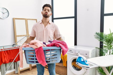Photo for Young man with beard holding laundry basket looking at the camera blowing a kiss being lovely and sexy. love expression. - Royalty Free Image
