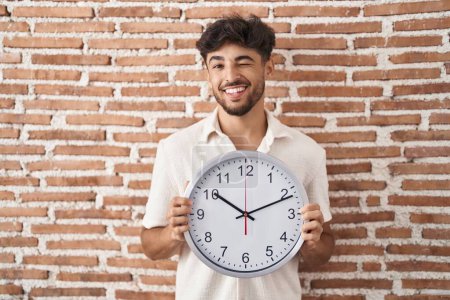 Foto de Hombre árabe con barba sosteniendo un gran reloj guiñando el ojo mirando a la cámara con expresión sexy, cara alegre y feliz. - Imagen libre de derechos
