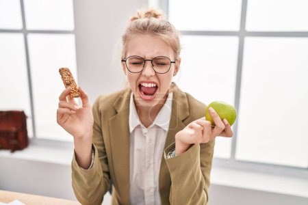 Photo for Young caucasian woman working at the office eating snack angry and mad screaming frustrated and furious, shouting with anger looking up. - Royalty Free Image