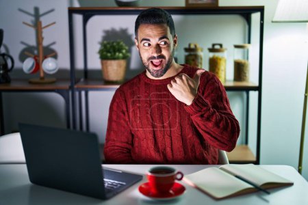 Foto de Young hispanic man with beard using computer laptop at night at home smiling with happy face looking and pointing to the side with thumb up. - Imagen libre de derechos