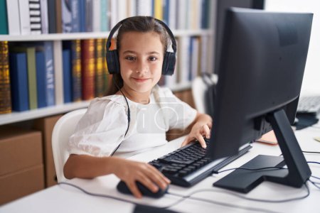 Téléchargez les photos : Adorable étudiante hispanique utilisant ordinateur et écouteurs assis sur la table en classe - en image libre de droit
