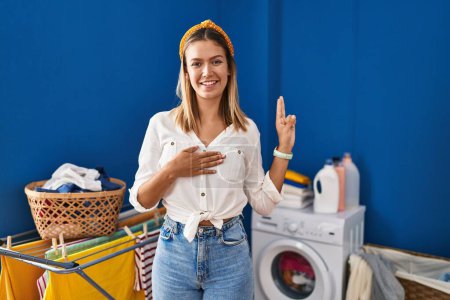 Téléchargez les photos : Young blonde woman at laundry room smiling swearing with hand on chest and fingers up, making a loyalty promise oath - en image libre de droit