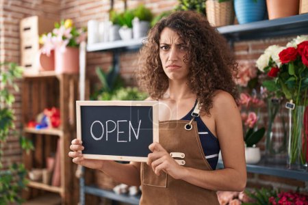 Photo for Hispanic woman with curly hair working at florist holding open sign clueless and confused expression. doubt concept. - Royalty Free Image