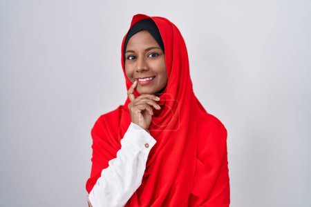 Téléchargez les photos : Jeune femme arabe portant l'écharpe traditionnelle islamique hijab regardant avec confiance la caméra souriant avec les bras croisés et la main levée sur le menton. penser positivement. - en image libre de droit