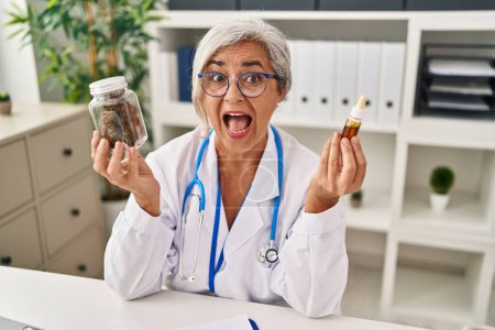 Foto de Middle age woman doctor holding cbd oil celebrating crazy and amazed for success with open eyes screaming excited. - Imagen libre de derechos