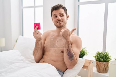 Téléchargez les photos : Young hispanic man holding condom in the bed pointing thumb up to the side smiling happy with open mouth - en image libre de droit