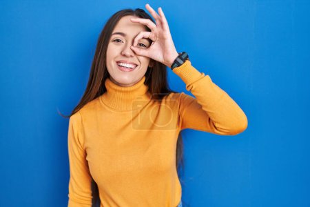 Photo for Young brunette woman standing over blue background doing ok gesture with hand smiling, eye looking through fingers with happy face. - Royalty Free Image