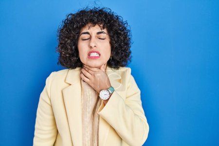 Foto de Young brunette woman with curly hair standing over blue background touching painful neck, sore throat for flu, clod and infection - Imagen libre de derechos