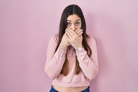 Foto de Young brunette woman standing over pink background shocked covering mouth with hands for mistake. secret concept. - Imagen libre de derechos