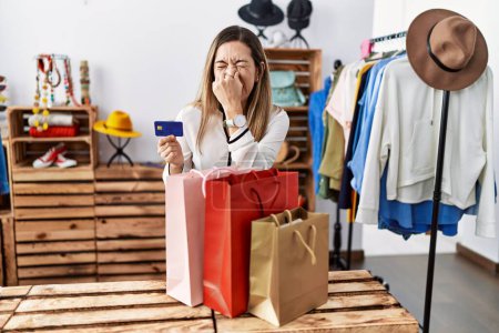 Foto de Mujer hispana joven sosteniendo bolsas de la compra y tarjeta de crédito en la tienda de ropa oliendo algo apestoso y repugnante, olor intolerable, conteniendo la respiración con los dedos en la nariz. mal olor - Imagen libre de derechos