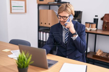 Photo for Young blond man business worker using laptop talking on smartphone at office - Royalty Free Image