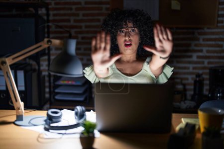 Foto de Young brunette woman with curly hair working at the office at night doing stop gesture with hands palms, angry and frustration expression - Imagen libre de derechos