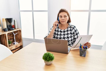 Foto de Mujer hispana de mediana edad trabajando en la oficina con portátil hablando por teléfono sonriendo mirando a un lado y mirando hacia otro pensando. - Imagen libre de derechos