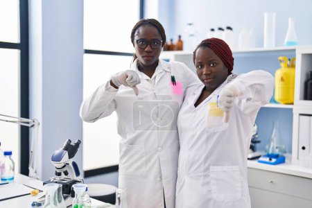 Foto de Dos mujeres africanas trabajando en el laboratorio científico con la cara enojada, signo negativo que muestra aversión con los pulgares hacia abajo, concepto de rechazo - Imagen libre de derechos