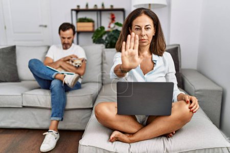 Foto de Hispanic middle age couple at home, woman using laptop doing stop sing with palm of the hand. warning expression with negative and serious gesture on the face. - Imagen libre de derechos