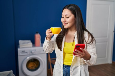 Photo for Young hispanic woman using smartphone and drinking coffee waiting for washing machine at laundry room - Royalty Free Image