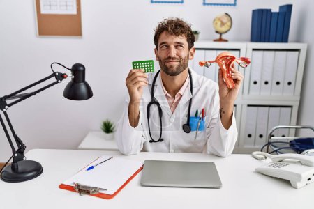 Photo for Young hispanic doctor man holding female genital organ and birth control pills smiling looking to the side and staring away thinking. - Royalty Free Image