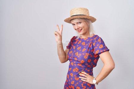 Téléchargez les photos : Jeune femme caucasienne portant une robe de fleurs et un chapeau d'été souriant regardant la caméra montrant des doigts faisant signe de victoire. numéro deux. - en image libre de droit