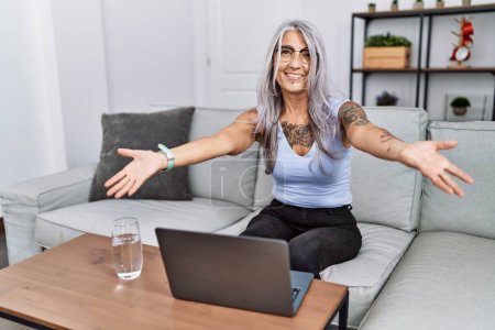 Téléchargez les photos : Femme aux cheveux gris d'âge moyen utilisant un ordinateur portable à la maison regardant la caméra souriant à bras ouverts pour se faire câliner. expression joyeuse embrassant le bonheur. - en image libre de droit