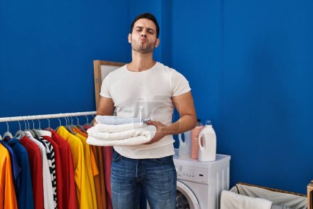 Foto de Young hispanic man with beard holding folded clean towels at laundry room looking at the camera blowing a kiss being lovely and sexy. love expression. - Imagen libre de derechos