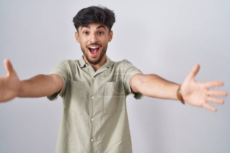 Téléchargez les photos : Arab man with beard standing over white background looking at the camera smiling with open arms for hug. cheerful expression embracing happiness. - en image libre de droit