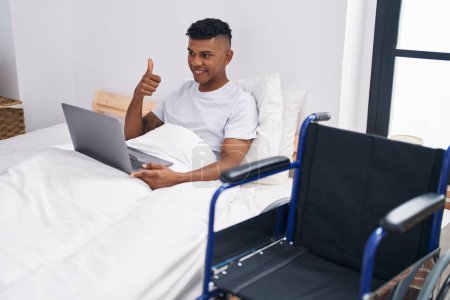Foto de Young hispanic man lying on the bed, using wheelchair smiling happy and positive, thumb up doing excellent and approval sign - Imagen libre de derechos