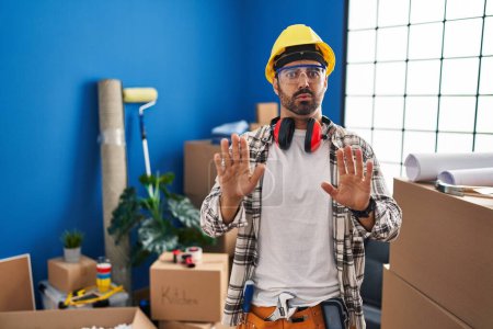 Foto de Young hispanic man with beard working at home renovation moving away hands palms showing refusal and denial with afraid and disgusting expression. stop and forbidden. - Imagen libre de derechos