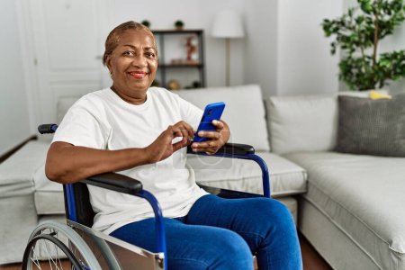 Foto de Mujer afroamericana mayor usando teléfono inteligente sentado en silla de ruedas en casa - Imagen libre de derechos