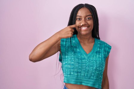 Foto de Young african american with braids standing over pink background pointing with hand finger to face and nose, smiling cheerful. beauty concept - Imagen libre de derechos
