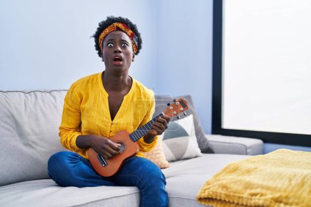 Foto de African young woman playing ukulele at home angry and mad screaming frustrated and furious, shouting with anger looking up. - Imagen libre de derechos
