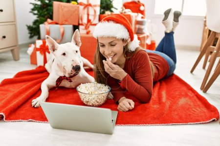 Foto de Young caucasian woman watching movie lying with dog by christmas tree at home - Imagen libre de derechos