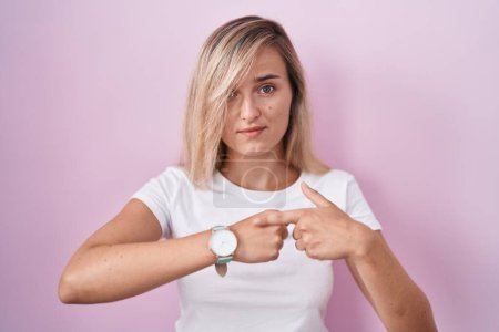 Téléchargez les photos : Young blonde woman standing over pink background in hurry pointing to watch time, impatience, looking at the camera with relaxed expression - en image libre de droit