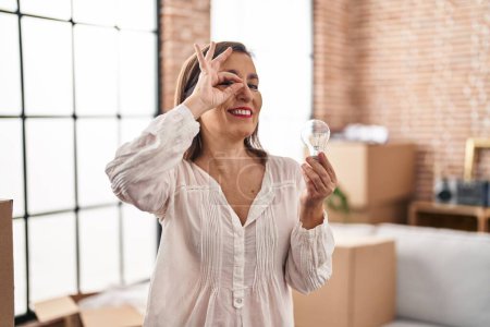 Foto de Middle age hispanic woman holding lightbulb for inspiration and idea smiling happy doing ok sign with hand on eye looking through fingers - Imagen libre de derechos