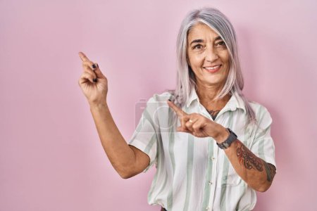 Photo for Middle age woman with grey hair standing over pink background smiling and looking at the camera pointing with two hands and fingers to the side. - Royalty Free Image