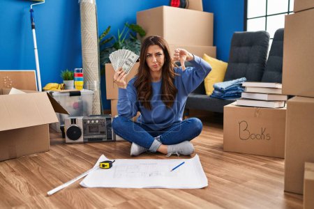 Photo for Young brunette woman sitting on the floor at new home holding money with angry face, negative sign showing dislike with thumbs down, rejection concept - Royalty Free Image