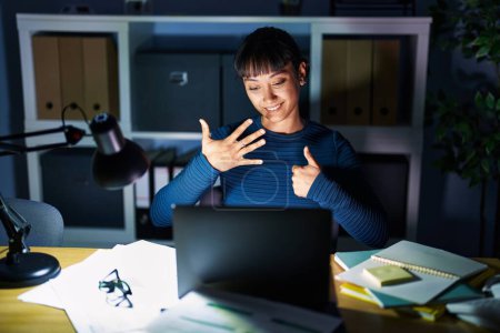 Foto de Young beautiful woman working at the office at night showing and pointing up with fingers number six while smiling confident and happy. - Imagen libre de derechos
