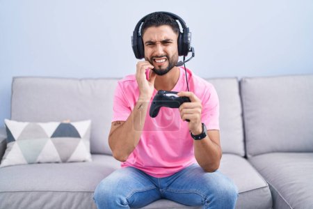 Téléchargez les photos : Hispanic young man playing video game holding controller sitting on the sofa touching mouth with hand with painful expression because of toothache or dental illness on teeth. dentist concept. - en image libre de droit