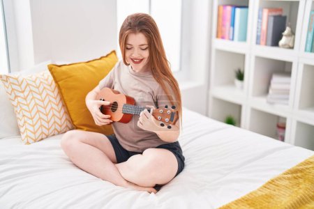 Foto de Joven pelirroja jugando ukelele sentado en la cama en el dormitorio - Imagen libre de derechos
