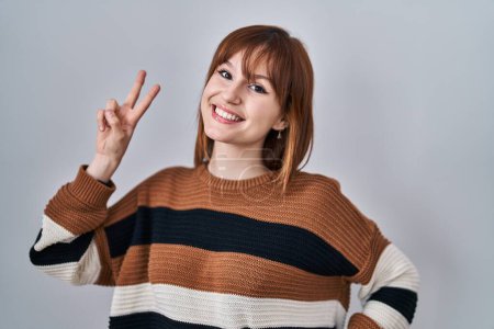 Téléchargez les photos : Young beautiful woman wearing striped sweater over isolated background smiling looking to the camera showing fingers doing victory sign. number two. - en image libre de droit