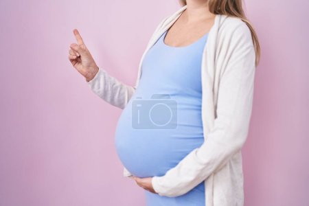 Foto de Mujer embarazada joven esperando un bebé, tocando el vientre embarazada sonriendo feliz señalando con la mano y el dedo a un lado - Imagen libre de derechos