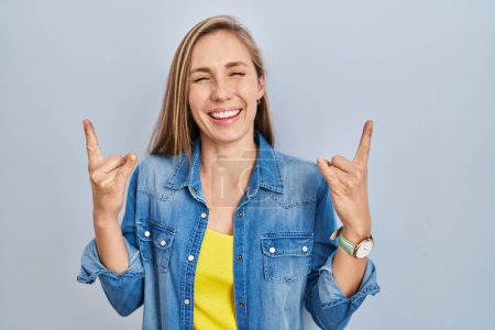 Photo for Young blonde woman standing over blue background shouting with crazy expression doing rock symbol with hands up. music star. heavy music concept. - Royalty Free Image
