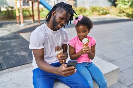 Foto de Father and daughter eating ice cream make selfie by smartphone at park - Imagen libre de derechos