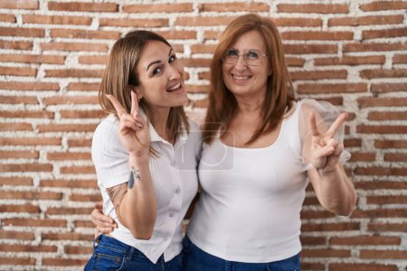 Téléchargez les photos : Hispanic mother and daughter wearing casual white t shirt smiling with happy face winking at the camera doing victory sign with fingers. number two. - en image libre de droit