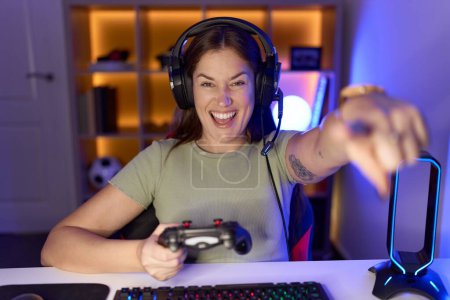 Foto de Hermosa mujer morena jugando videojuegos con auriculares que apuntan a usted y la cámara con los dedos, sonriendo positiva y alegre - Imagen libre de derechos