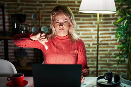 Foto de Blonde woman using laptop at night at home looking unhappy and angry showing rejection and negative with thumbs down gesture. bad expression. - Imagen libre de derechos