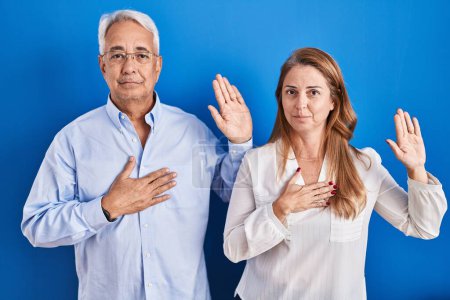 Foto de Middle age hispanic couple standing over blue background swearing with hand on chest and open palm, making a loyalty promise oath - Imagen libre de derechos